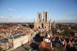 Aviw of Castle square - which is not a square at all. In the foreground is the cobbled surface of the open space which is surrounded by historic buildings. From left to right, the regency assembly rooms, a Georgian 3 story house, then Leigh-Pemberton house: a half-timbered building with the upper story jettied out. Then a medieval church and the entrance to the Cathedral Close. In the left distance the towers of the cathedral rise into a blue sky.