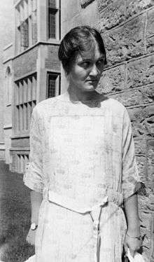 A woman in a white dress stands in front of a stone university building and looks down and to one side.