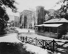 Front of the arsenal, viewed at sharp angle, with small building and rustic fence in foreground, next to it