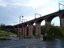 a viaduct consisting of a series of arches made of red brick, with a road passing under it and trees and a grass bank behind it