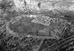 Large circular depression outlined by a stone wall. The bottom is flat and grassy, and has a collection of rectangular stone foundations and smaller circles of stone. A great sandstone cliff towers in the background, and beneath the cliff are other stone foundations that are larger and higher.