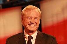 A smiling man with blond hair wearing a gray suit jacket and black tie.