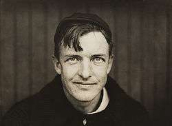 A dark-haired man wearing a black sweater and crownless baseball cap looks into the camera. His hair hangs down over his right side of his forehead, and he has a slight smirk on his face.