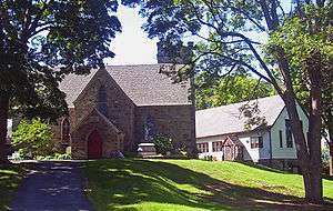 A stone building, shaded by tall trees, with a small pointed section projecting from the front, which has another small pointy-roofed section with red door. There is a tower on the back of the building and a white house-like building to the right.