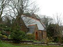 A small church seen from the southwest with a south porch. At the far end a saddleback-roofed tower is just visible.  Around the church are trees and bushes and in the foreground is a wall