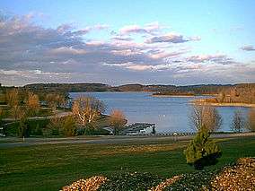 A blue lake with a dock in rolling green hills, under a partly cloudy sky