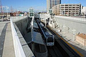 A white train is parked at a recessed side platformed station with canopies and signage visible.