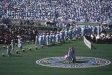 A bunch of people standing with cap and gowns while two people stand on a grass field.