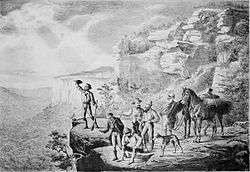 Image shows the group of explorers standing at the edge of a cliff, looking out over plains.