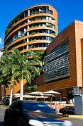 Tall, round light-brown building with palm trees in front and a car passing