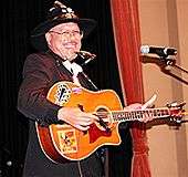 A man wearing black clothing and a black hat, holding a guitar and standing behind a microphone