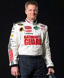 A man in his thirties wearing a white jacket with sponsors logos. He is holding a crash helmet in his left hand.