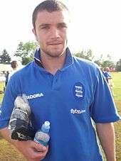 An unshaven white man in a blue sports shirt stands on a grassy area.