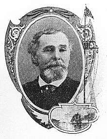 Head of a white man with a bushy goatee and thinning hair, wearing a dark suit coat and tie.