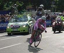 A cyclist on his bicycle, wearing a skintight pink jersey with pink shorts and shoes. A lime green car and two motorcycles follow behind him, and spectators are visible on the roadside behind barricades.