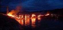 Fire consuming a bridge, with portions of the deck falling to the river below.