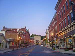 A view of an urban street at dusk with brick buildings on either side and a brown building with a large arched window in the distance