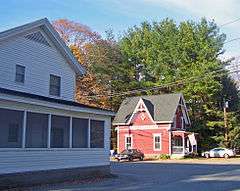 A red house with decorative vergeboards at right in the distance in front of some trees. A white house at left is closer to the camera and fills that side of the image.