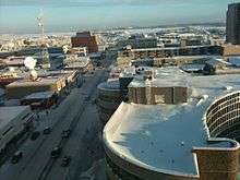 Snow-covered flat roofs and snowy streets seen from slightly above. In the far background is a frozen lake and some low hills