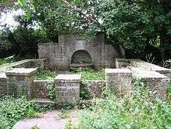 Stone wall with arched gap, overgrown with weeds.