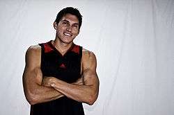 A muscular man wearing a black tanktop with red on the shoulder straps which his arms crossed over his upper abdomen.