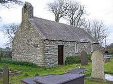 A small plain stone church seen from an angle with a bellcote on the near gable, and a simple door and two windows along the side