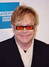 A Caucasian man with light brown hair. He is wearing black framed glasses with orange lenses and a black suit over a white shirt. Behind him is a whit wall with a blue square.