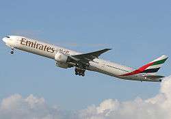 A mostly white Boeing 777, with some red, green and black markings, of Emirates, in flight, facing left.