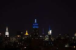 Upper floors of the Empire State Building lit in blue, amid other lit skyscrapers