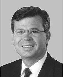 A man with dark hair and glasses wearing a black jacket and tie and white shirt