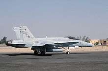 Photo of a modern fighter aircraft moving along tarmac. Sand and buildings are visible in the background.