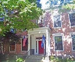 A brick building with a red door behind a small columned porch with the U.S. and New York flags behind a large tree on the left