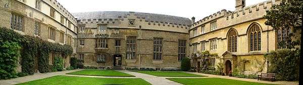 Adjoining buildings (all with battlements) on three sides of an area of grass in the middle of a quadrangle crossed by pathways from side to side and front to back; at the far side, a large building with a clock at roof level and three large windows; on the left, regularly placed windows in a three-storey building; on the right, a chapel with three arched windows alongside a three-storey house