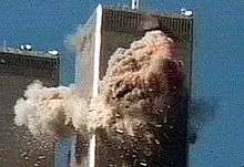 A high-rise tower covered by debris on two of its faces. In the lower left corner is a similar building.