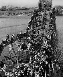 Black-and-white photo of people crossing a river via a destroyed bridge