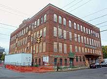 A four-story square brick building with boarded-up windows. There is a white trailer on its left and some signs from a construction company, both in a fenced-off area