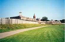 Photograph of a grassy field and a log stockade. The tops of log buildings are visible behind the stockade.