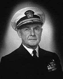 Head and shoulders of man in dark suit and tie wearing white peaked cap with gold braid. He has several rows of ribbons on the left breast.