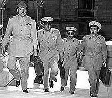 Four men in military uniform, wearing buttoned jackets, wide-rimmed hats, black shoes, and carrying briefcases. They are ascending the stairs of a building.