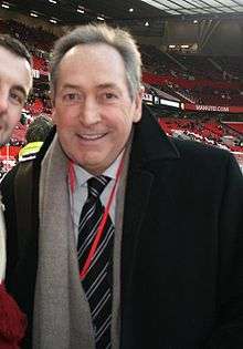 The upper body of a grey haired man. He is wearing a black coat, white shirt, black tie with blue stripes, grey scarf and a red ribbon is visible around his neck.