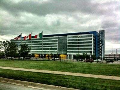 A wide rectilinear six-story blue-and-white building with the American, Canadian and Mexican flags flying in front, seen from a nearby roadway, under a cloudy sky