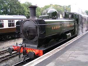 A green pannier tank locomotive standing at a platform with a red passenger carriage behind. The lettering "GREAT WESTERN" is shown in yellow on the side of the pannier tank.