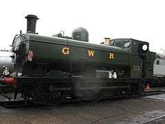 A pannier tank locomotive is standing over an inspection pit. The locomotive is mainly painted green above the running plate, although the chimney is black and the safety valve cover is polished brass. The letters "G W R" are shown in yellow with red shading on the side of the pannier tank. The front buffer beam is painted red.