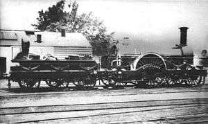 A black and white image showing a steam locomotive facing to the right. The low tender on the left has six wheels; the engine itself has a large wheel in the middle with two wheels in front and one behind.