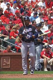 A man wearing a black baseball jersey with "Mets" written across the chest, gray pants, and a black batting helmet holds his baseball bat as he approaches home plate to begin his at-bat.