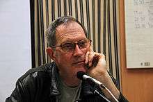 Head and shoulders of a man wearing a green t-shirt and black leather jacket. His hair is thinly cropped looks like he's contemplating something.