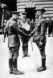 Two men in military uniform smiling and shaking hands. The man on the left is older and slightly stooped. People are gathered in the background.