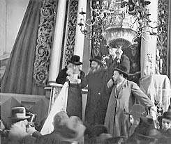 A sepia photograph shows a rabbi standing on the synagogue podium, addressing the congregation. He is reading from a sheet of paper, and three other men stand to his left. Behind him can be seen the white columns and ornate carvings of the holy ark. A chandelier, suspended from the ceiling, appears at the top right.