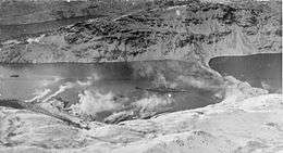 Black and white photograph of a World War II-era warship in a small bay with a steep and snow-covered shoreline. Clouds of smoke are being blown from the shoreline towards the ship.