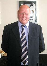 Portrait photograph of a smiling bald man wearing a black jacket over a blue shirt and striped tie.